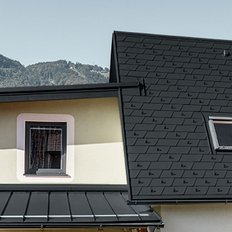 Das Dach eines Hauses mit schwarzen Schindeln, einer kleinen Dachgaube und einem Schornstein, umgeben von grünen Bergen.