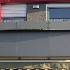 Ein grauer Balkon mit Pflanzen steht vor einer roten Wand, während ein Sonnenschutz darunter heruntergelassen ist.