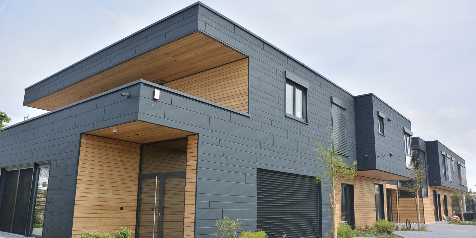 Moderne Wohnhäuser mit einer Kombination aus dunkler Fassade und Holzverkleidung, umgeben von kleinen Pflanzen und Bäumen.