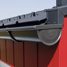 Ein modernes Dach mit einer schwarzen Dachrinne, die an einer leuchtend roten Wand befestigt ist, unter klarem Himmel.